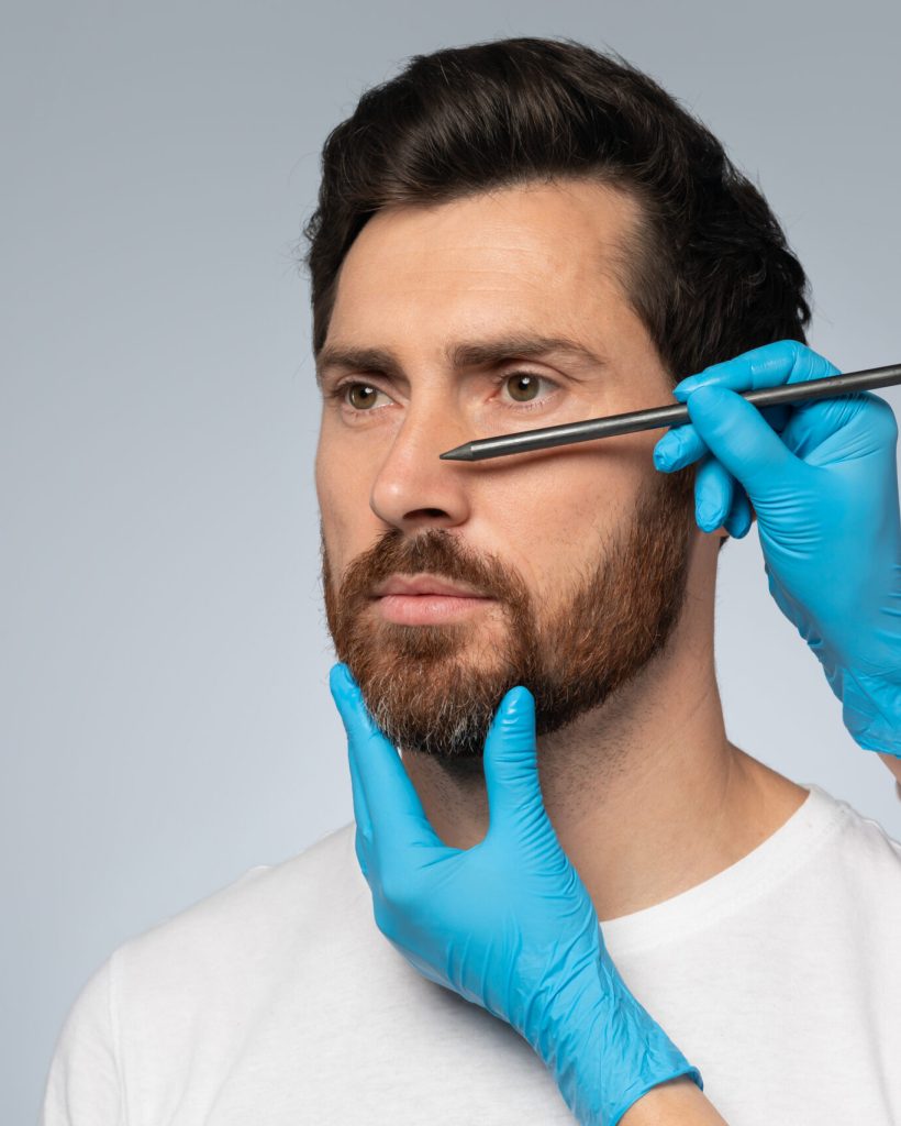 Portrait of middle aged man getting prepared for plastic surgery procedures by doctor drawing contours on his nose, standing on grey background, copy space